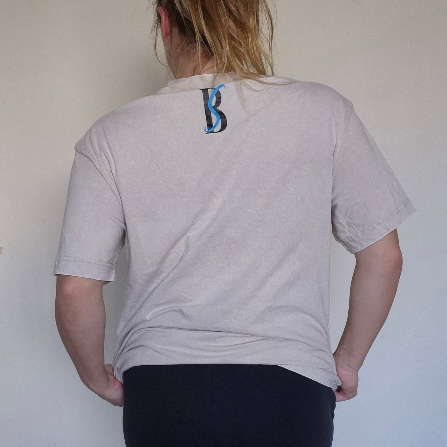 Person wearing a light gray t-shirt with a logo on the back, standing against a plain white wall.