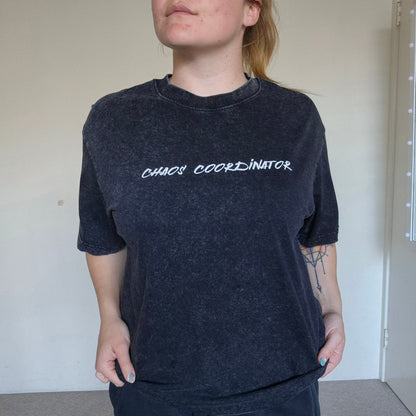 Person wearing a black t-shirt with 'CHAOS COORDINATOR' text in a plain room.